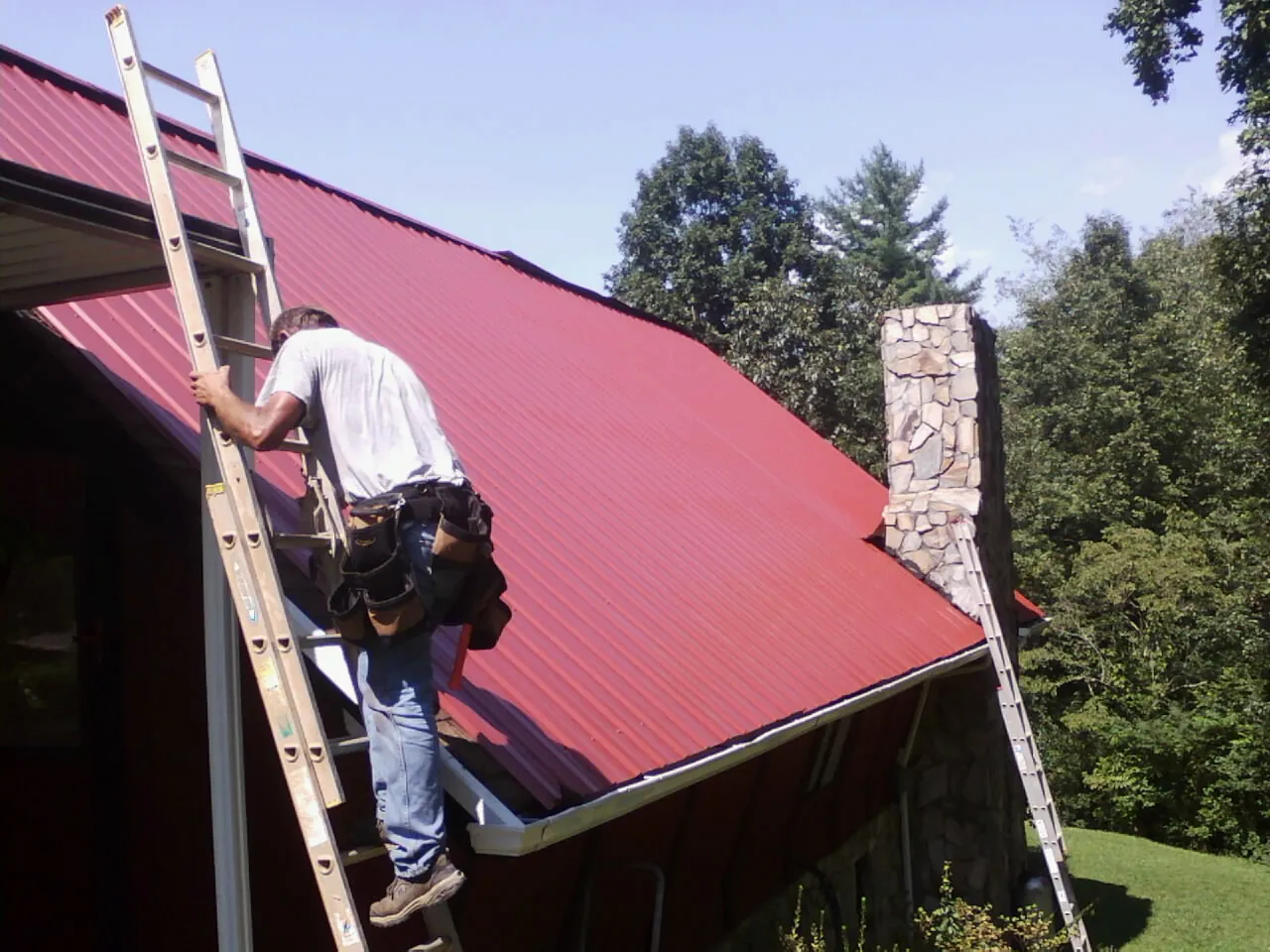 Roofer on a ladder beside a red metal roof and stone chimney — Soffit & Fascia Repair by Roofing Brothers in Vian